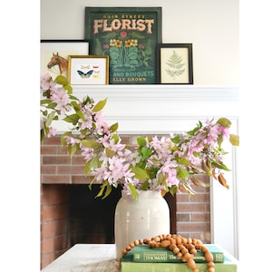 May include: A white ceramic vase with a pink and green floral arrangement on a wooden table. The vase is decorated with a rustic, distressed finish. The flowers are arranged in a cascading style, with the stems extending over the edge of the vase. The table is decorated with a stack of books and a wooden bead garland.