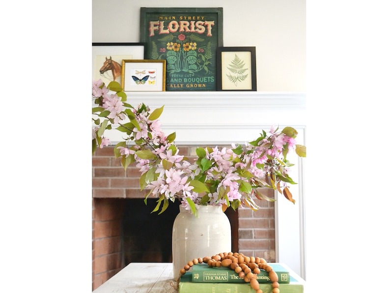 May include: A white ceramic vase with a pink and green floral arrangement on a wooden table. The vase is decorated with a rustic, distressed finish. The flowers are arranged in a cascading style, with the stems extending over the edge of the vase. The table is decorated with a stack of books and a wooden bead garland.