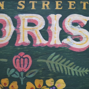 May include: A vintage-style sign with the words "Main Street Florist" painted in yellow and white letters on a pink background. The sign has a green wooden background with painted floral designs.