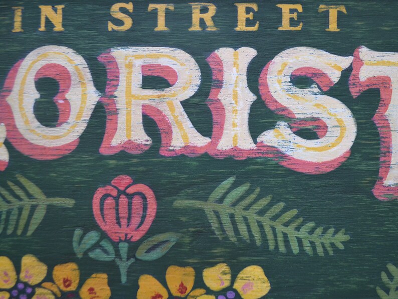 May include: A vintage-style sign with the words "Main Street Florist" painted in yellow and white letters on a pink background. The sign has a green wooden background with painted floral designs.