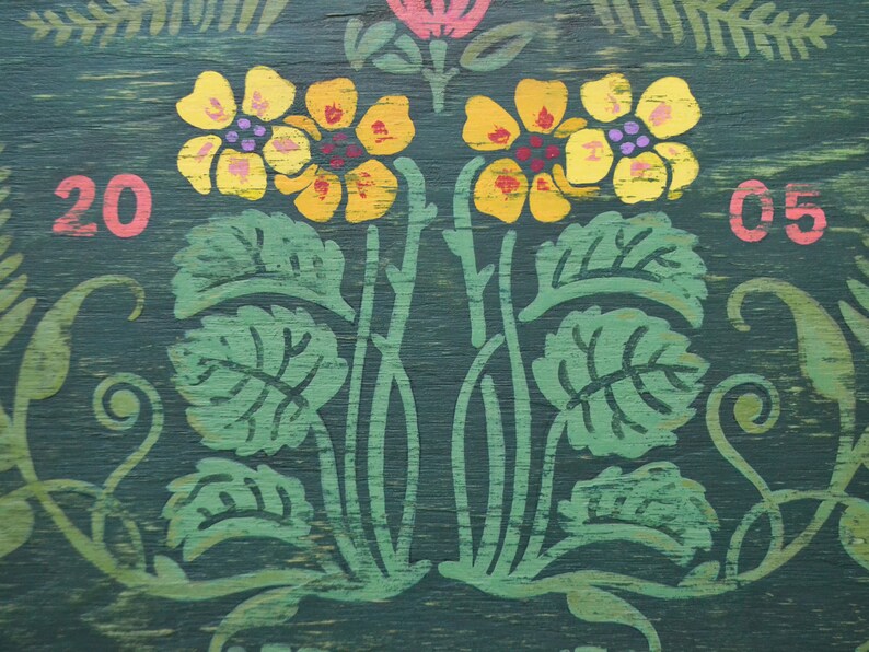 May include: A green wooden sign with yellow flowers and green leaves painted on it. The numbers 20 and 05 are painted in pink on either side of the flowers.