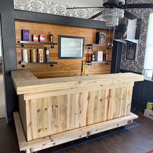 May include: A custom-built wooden bar with a light-colored finish. The bar features a countertop, a front-facing structure, and a back wall with shelves. The back wall has a mirror with the text "THE ROB ROY". The bar is on wheels.