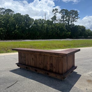 May include: A large L-shaped wooden bar counter featuring a dark brown stained finish with visible wood grain. The structure has a flat top surface, vertical paneling on the front, and is mounted on small wheels for mobility on the ground.