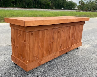 May include: A wooden bar with a brown stain finish. The bar has a flat top and is on wheels. The bar is in front of a grassy area with a railway track in the background.