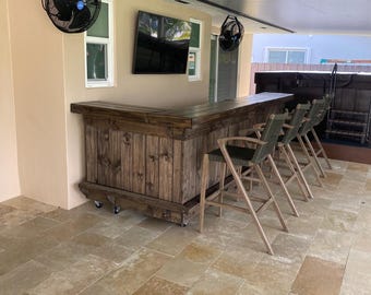 May include: A wooden bar with a rustic finish, featuring a built-in countertop and a curved design. The bar is equipped with wheels for easy mobility and is accompanied by four bar stools with green cushions. The bar is situated on a tiled patio with a ceiling fan overhead.