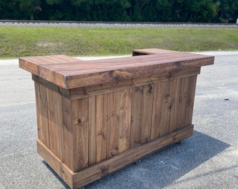 May include: A wooden bar with a dark brown stain, featuring a rectangular top and a curved front. The bar is on wheels and has a rustic, farmhouse style.