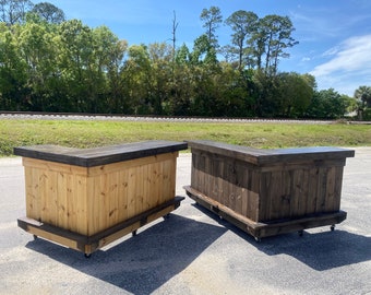 May include: Two wooden bar carts with a dark brown stained top and a natural wood finish on the sides. The bar carts are on wheels and have a corner design.