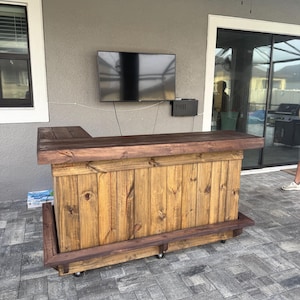 May include: A wooden outdoor bar with a stained finish. The bar has a countertop, a footrest, and is mounted on wheels. The bar is made of vertical wooden planks and is set against a gray wall with a mounted TV.