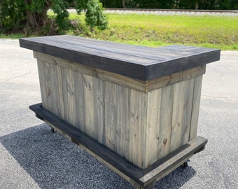 May include: A grey wooden bar with a black stained top and black trim. The bar is on wheels and has a rustic look.