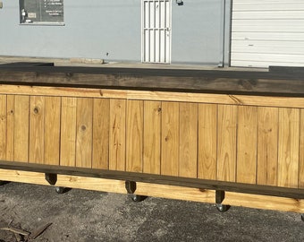 May include: A wooden bar counter with a dark brown top and a light brown base. The counter has a built-in shelf and is on wheels.