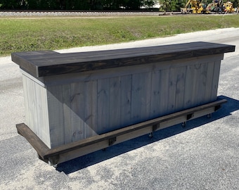May include: A gray wooden bar with a dark stained top and footrest. The bar is constructed from vertical planks and has a rectangular shape with a small extension on one side. It sits on small wheels.