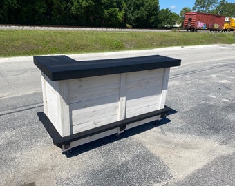 May include: A white wooden bar with a black top and black trim. The bar is on wheels and has a shelf underneath.