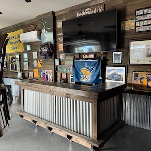 May include: A custom-built bar with a wooden frame and corrugated metal siding. The bar has a dark stained countertop and is decorated with sports memorabilia and a television. The bar is on wheels.