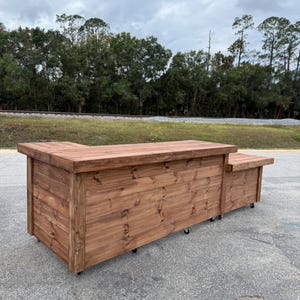 May include: A large, rectangular wooden bar with a rich brown stain. The bar has a countertop and a lower section, both constructed from horizontal planks. It sits on small black wheels for mobility. The background features trees and a cloudy sky.