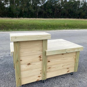 May include: A wooden counter with a light wood finish and a natural wood grain. The counter has a rectangular top and a smaller rectangular shelf underneath. The counter is on wheels and has a light green trim.
