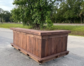 May include: A dark brown wooden bar with a corner design, mounted on wheels. The bar is made of wood planks and has a simple, rustic design.