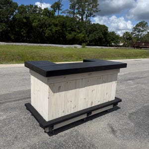 May include: A wooden bar with a black top and base, and a natural wood finish on the front. The bar is rectangular and appears to be on wheels. The bar is outdoors with trees and a blue sky in the background.