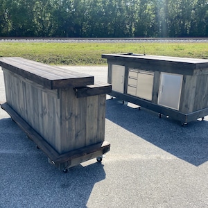 May include: Two wooden outdoor kitchen islands with a gray stained finish. The island on the left has a bar top and is on wheels. The island on the right has a stainless steel sink and drawers and is also on wheels.