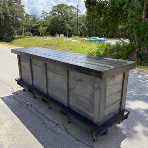May include: A large, rectangular, wooden bar with a dark gray stain. The bar has a flat top and is mounted on wheels for easy mobility. The bar is constructed from wooden planks and is suitable for indoor or outdoor use.