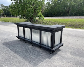 May include: A black wooden bar with a corrugated metal back panel and wheels. The bar has a black countertop and is made of reclaimed wood.