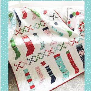 May include: A white quilt with a pattern of Christmas stockings in various colors, including red, green, and blue. The quilt has a red border and the text "Pixie Stockings by tasha noel".