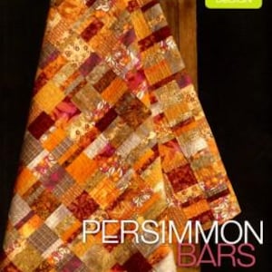 May include: A Persimmon Bars quilt with a patchwork design in shades of orange, brown, and burgundy. The quilt is draped over a wooden surface. The text "PERSIMMON BARS" is displayed in white letters.