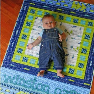 Puede incluir: Un bebé yace sobre un edredón colorido con un diseño azul y verde. El edredón presenta un patrón de coches y aviones, con el nombre "Winston Ozro" bordado. El edredón es parte de la colección "Lullaby Lane" de Sassafras Lane Designs.