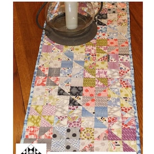 Könnte beinhalten: Ein Patchwork-Tischläufer, 35 x 89 cm, mit einem quadratischen Charm-Design. Der Läufer weist eine Vielzahl von Stoffmustern und -farben auf, darunter Blumenmuster, Punkte und geometrische Formen. Der Text "Square Cut Charm" und "Creek Side Stitches" ist sichtbar.