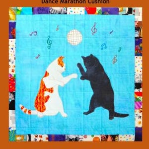 May include: A square cushion with a blue center featuring two dancing cats, one white and orange, the other black, under a disco ball and musical notes. The cushion is bordered by a patchwork design, with the text "Trouble & Boo Designs" and "Dance Marathon Cushion".