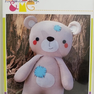 May include: A plush teddy bear with a light pink body, blue patches, and a white belly. The bear has black button eyes, a stitched black nose, and red cheeks. The word "BUDDY" is printed at the bottom of the image.
