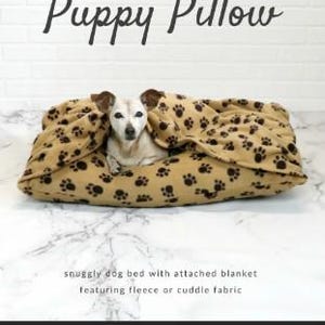 May include: A tan dog bed with a paw print pattern and attached blanket. The text "Puppy Pillow" is at the top, with "snuggly dog bed with attached blanket featuring fleece or cuddle fabric" below. The Sallie Tomato logo is at the bottom.
