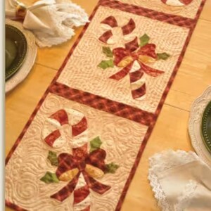 May include: A quilted Christmas table runner featuring candy cane designs. The runner has a cream-colored background with red and white candy canes, red bows, and green holly leaves. The edges are trimmed with a red plaid pattern.