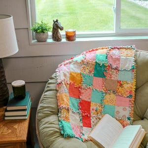 Könnte beinhalten: Eine bunte Patchwork-Decke mit Rüschenkanten liegt auf einem grünen Papasan-Stuhl. Die Decke zeigt Quadrate aus verschiedenen floralen und unifarbenen Stoffen in Rosa, Gelb und Türkis. Ein offenes Buch liegt auf dem Stuhl, mit einem Fenster und einer grünen Wiese im Hintergrund.
