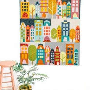May include: A colorful, patchwork quilt featuring a whimsical village scene. The quilt showcases rows of stylized buildings in various colors, including yellow, blue, and red, with trees interspersed. The background is a light gray with a dotted pattern.