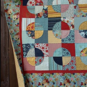 Puede incluir: Un edredón colorido con un diseño de patchwork que presenta patrones de cuarto de círculo en varias telas. El edredón tiene un borde floral y el texto "Broken Coins".