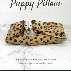 Puede incluir: Una cama para perros de color beige con un estampado de huellas de patas y una manta adjunta. El texto "Puppy Pillow" se muestra encima de la cama. La cama está hecha de forro polar o tela suave. El texto "Sallie Tomato" se muestra en la parte inferior.