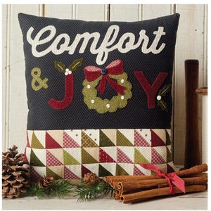 May include: A decorative square pillow with the words "Comfort & JOY" in white and red appliqued letters. The pillow features a quilted design with a Christmas wreath and a patchwork border of triangles in red, green, and beige. Cinnamon sticks and pine cones are in the foreground.