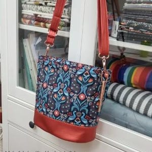 May include: An oval-shaped bag with a floral pattern in blue, teal, and orange, featuring a red base and straps. The bag measures 10" wide, 9" high, and 6.5" deep. The text "The Awesome Oval Bag" is at the top.