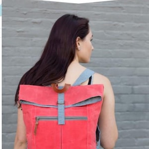 Pode incluir: Uma mochila de cor coral com aba, com um bolso frontal com zíper e uma alça xadrez azul e branca. A mochila é feita pela Noodlehead.