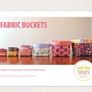 May include: Six fabric buckets of different sizes, each with a unique patterned fabric exterior. The text "FABRIC BUCKETS" is displayed in pink. The image also includes the text "A modern sewing pattern by Tamara Kate Design".