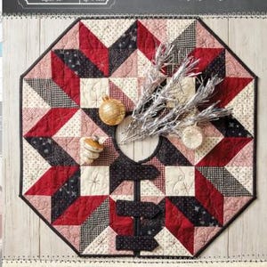 Könnte beinhalten: Ein gesteppter Weihnachtsbaumrock mit Sternmuster in Rot, Schwarz und Creme. Der Rock ist achteckig, mit einer zentralen Öffnung und dekorativen Ornamenten. Der Text "Christmas at Buttermilk Acres Tree Skirt" ist sichtbar. Größe: 61 x 61 cm.