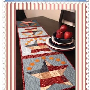 Puede incluir: Un camino de mesa patriótico de patchwork con diseños de estrellas y rayas en rojo, blanco y azul. El camino de mesa está sobre una mesa de madera oscura, con un cuenco de manzanas rojas y platos en el fondo. El texto "Shabby Fabrics" está en la parte superior.