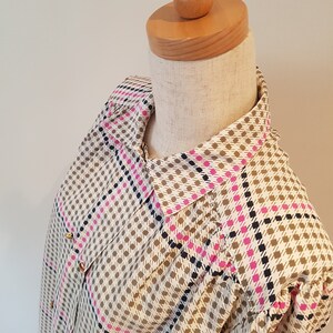 May include: A patterned button-down shirt displayed on a mannequin. The shirt features a houndstooth design in beige, brown, pink, and navy blue. Gold-colored buttons accent the front. The collar and sleeves are visible.