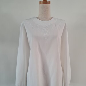 May include: White long-sleeve blouse with intricate embroidered detailing around the neckline. The blouse is displayed on a mannequin, showcasing its classic design and tailored fit. The embroidery features a delicate, swirling pattern.