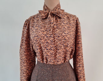 May include: A brown and blue geometric patterned blouse with a bow tie. The blouse has long sleeves and a high collar. The blouse is on a mannequin and is paired with a brown tweed skirt.