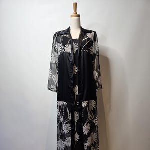 Könnte beinhalten: Ein zweiteiliges Kleid und Jacke mit schwarz-weißem Blumenmuster auf einer Schaufensterpuppe. Das Midikleid hat ein schwarzes Oberteil und ein weißes Blumenmuster auf dem Rock. Die Jacke ist transparent mit weißem Blumendesign.