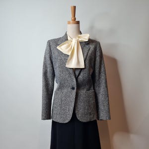 Puede incluir: Una chaqueta de tweed gris con un cierre de botón único y una falda negra. La chaqueta tiene una corbata de lazo de satén blanco.