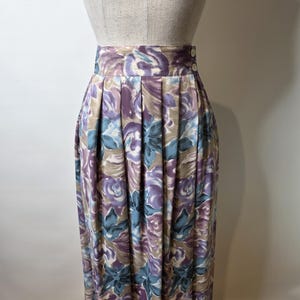 May include: A vintage midi skirt with a floral pattern in shades of purple, blue, and beige. The skirt features pleats and a button closure. The skirt is displayed on a mannequin.
