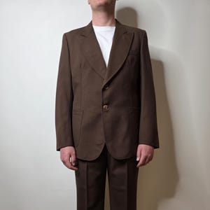 May include: A solid brown suit consisting of a two-button blazer with wide lapels and matching trousers. The blazer features two front pockets and is worn over a plain white t-shirt. The trousers have a straight-leg cut with a visible center crease.