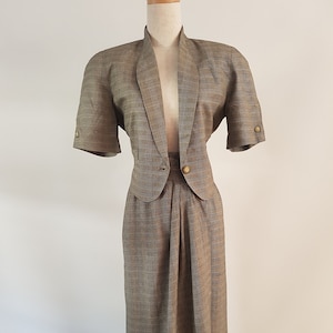 May include: A brown and gray plaid skirt suit with a short-sleeved jacket and a knee-length skirt. The jacket has a collar and a single button closure. The skirt has a waistband and a pleated front.